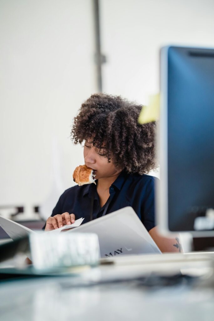 A dedicated woman multitasking with documents and food in a modern office setting.