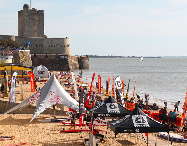 Stands, drapeaux et participants réunis pour un événement nautique sur une plage au pied d’un fort.
