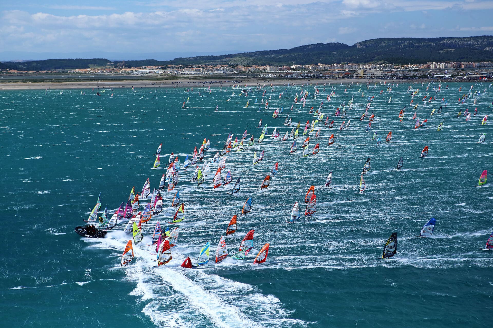 Grande flotte de pratiquants de windsurf aux voiles colorées naviguant en mer près de la côte.