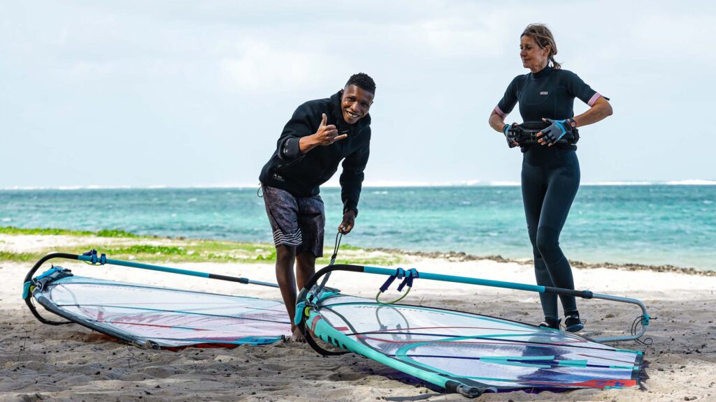Deux personnes en tenue de navigation sur une plage, près de voiles de planche à voile posées sur le sable.