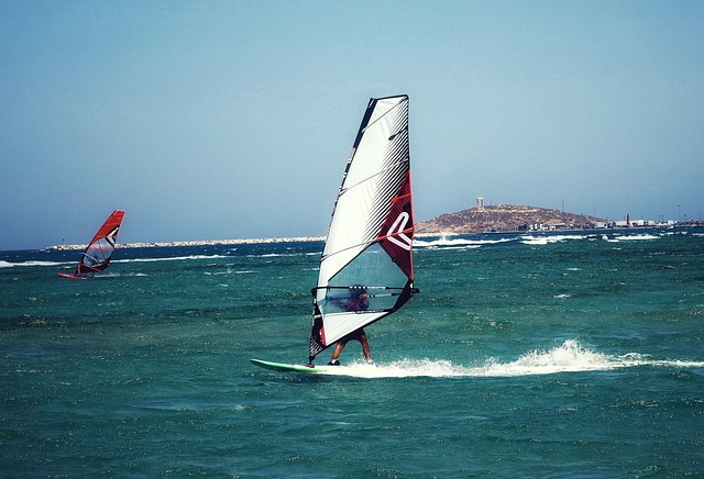 Deux personnes pratiquent la planche à voile sur une mer turquoise, sous un ciel bleu dégagé. Les voiles blanches et rouges contrastent avec les couleurs de l’eau. À l’arrière-plan, on aperçoit la côte et une île. L’image évoque les loisirs nautiques, le vent, la liberté et les vacances ensoleillées.